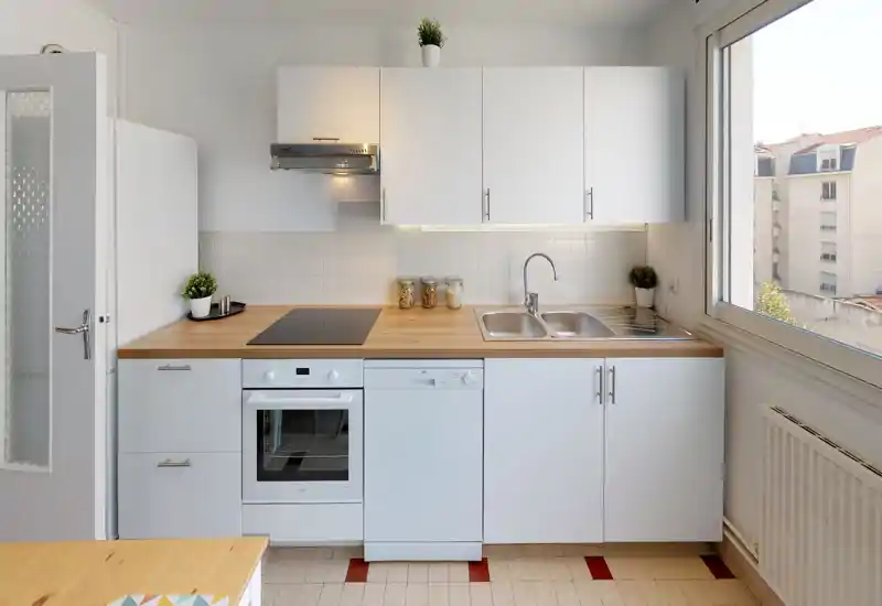 Cucina luminosa e minimal con pensili bianchi, piano di lavoro in legno, forno e piano cottura integrati, doppio lavello e ampia finestra che illumina l'ambiente.