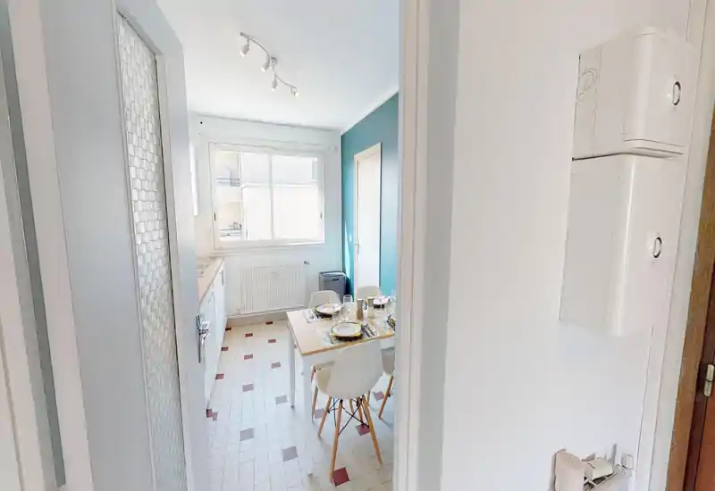 Cucina compatta e luminosa con un piccolo tavolo da pranzo per quattro persone. La grande finestra garantisce luce naturale e la parete azzurra dona un tocco moderno.