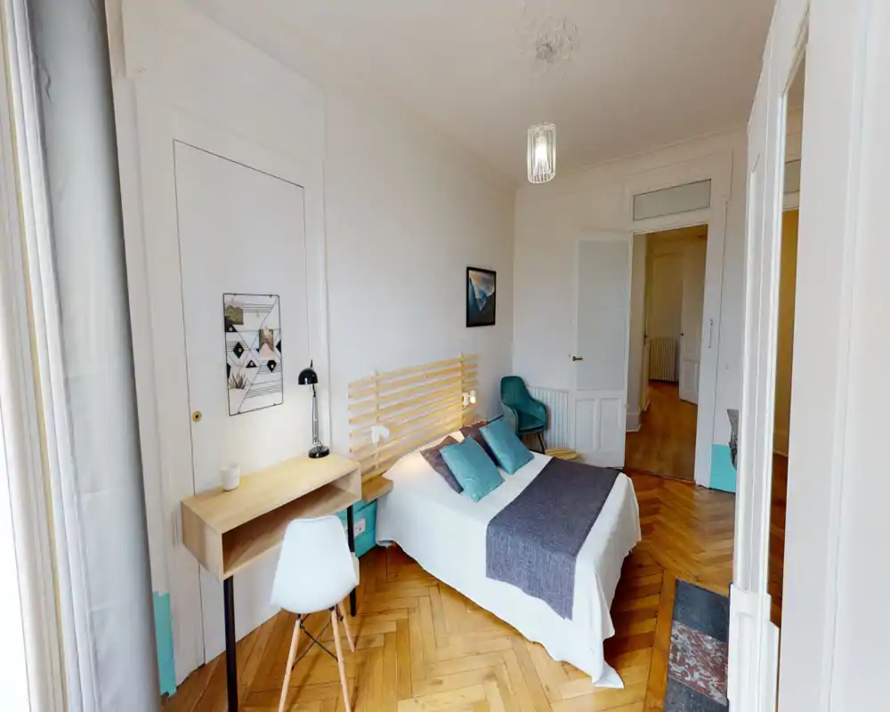 Camera da letto luminosa e ben arredata con letto matrimoniale centrale, testiera in doghe di legno, cuscini accentuati turchesi e una piccola scrivania vicino alla finestra — pavimento in parquet caldo e luce naturale morbida.