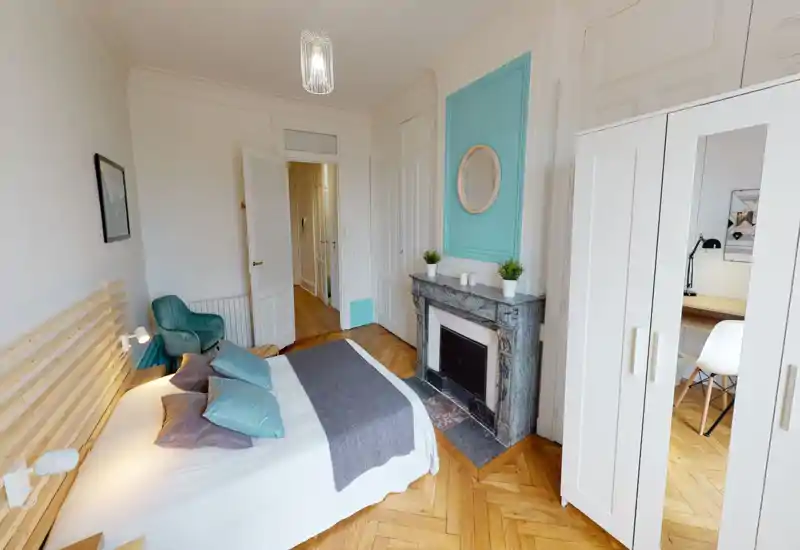 Camera da letto luminosa e di stile con un letto ampio, cuscini decorativi, una poltrona accogliente e un caminetto classico. Pareti neutre e accenti turchesi creano un'atmosfera moderna e rilassante.