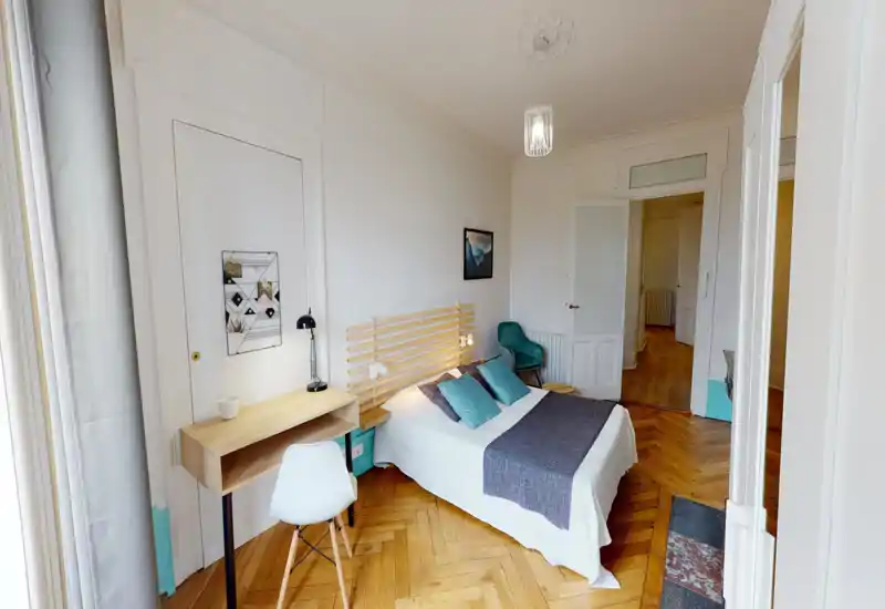 Camera da letto luminosa e ben arredata con letto matrimoniale centrale, testiera in doghe di legno, cuscini accentuati turchesi e una piccola scrivania vicino alla finestra — pavimento in parquet caldo e luce naturale morbida.