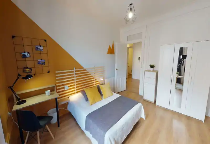Bright, stylish bedroom with a double bed, wooden slatted headboard and warm mustard-accent wall. The room includes a small desk, wardrobe with mirrored door and light wooden flooring — a cozy, well-lit space for relaxing or working.