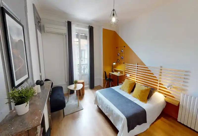 Bright, stylish bedroom with a double bed, wooden slatted headboard and warm mustard accent wall. Natural light from a tall window, bedside lamps and a small workspace make this room comfortable for rest and remote work.