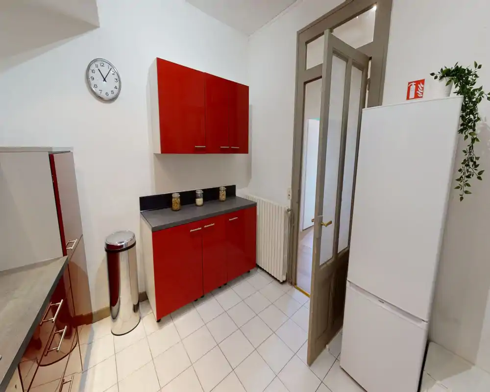 Clean modern kitchen with glossy red cabinets, grey countertops and a freestanding white fridge; bright tiled floor and simple decorative jars create a neat, functional cooking area.
