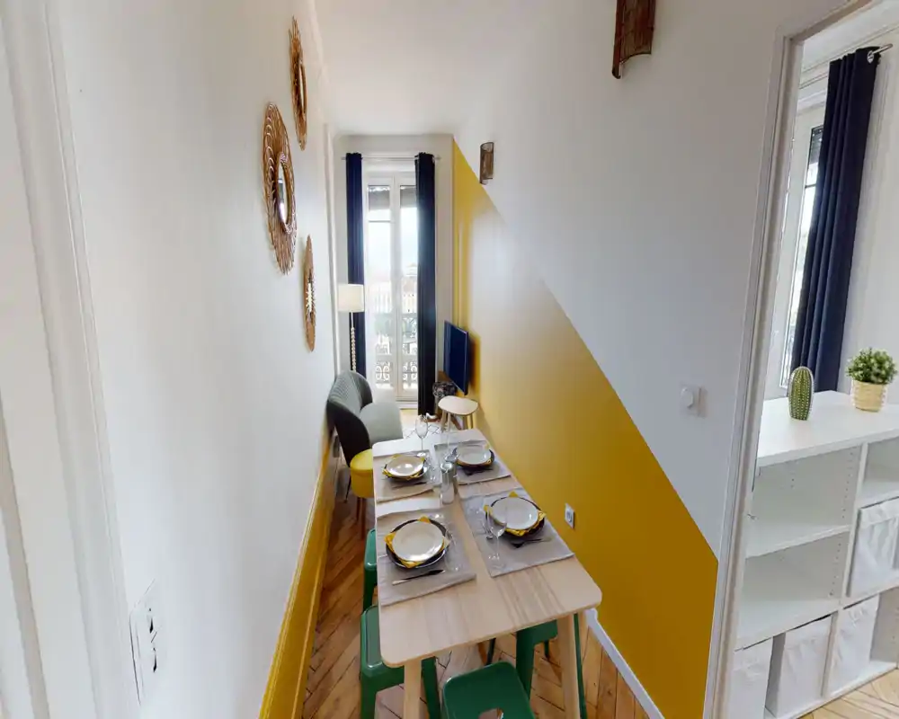 Narrow, stylish living area with a small dining table set for four, a cozy armchair, and a wall-mounted TV facing a balcony window. Warm yellow accent wall, decorative mirrors and natural light create an inviting atmosphere.