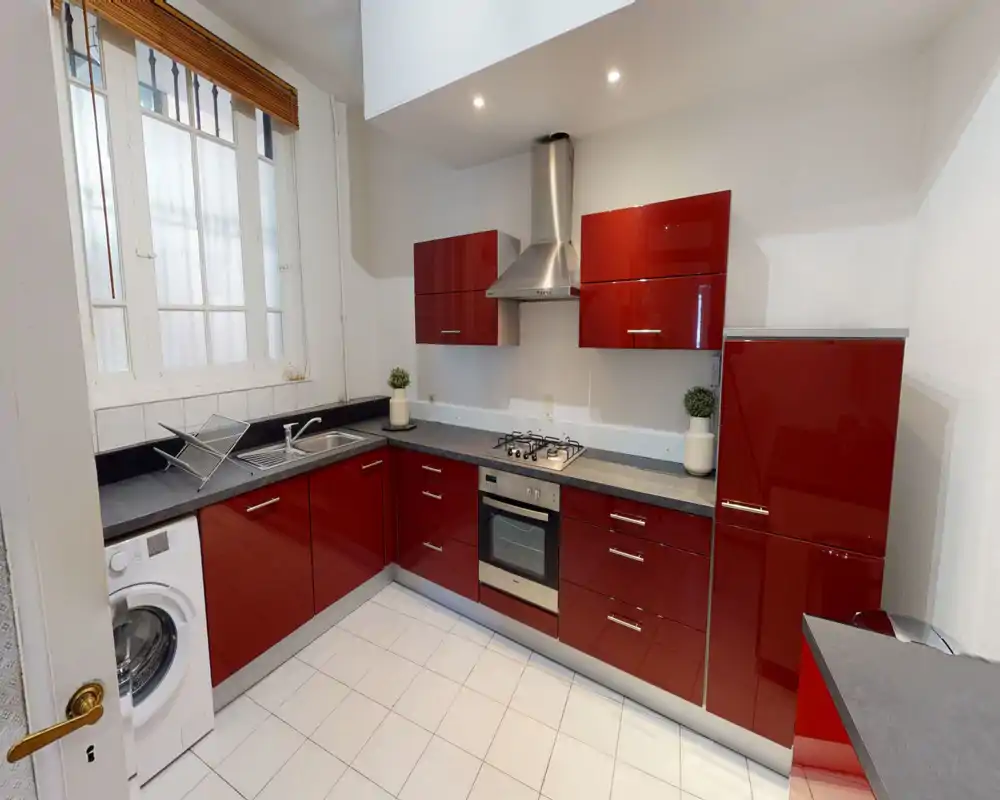 Bright modern kitchen with glossy red cabinets, built-in oven and gas hob, stainless extractor hood and a sink next to a large window. Clean tiled floor and compact layout ideal for efficient cooking.