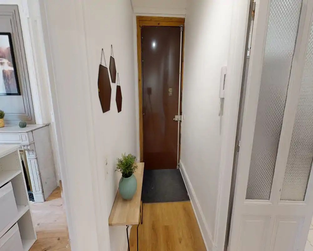 Small, tidy entrance hallway with a wooden bench, decorative vase and wall-mounted mirrors, leading to the main door. Clean white walls and a visible intercom make it practical for arrivals.