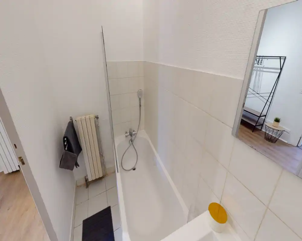 Compact bathroom with a tiled bathtub, handheld shower and radiator; neutral white walls and simple accessories create a clean, practical space.