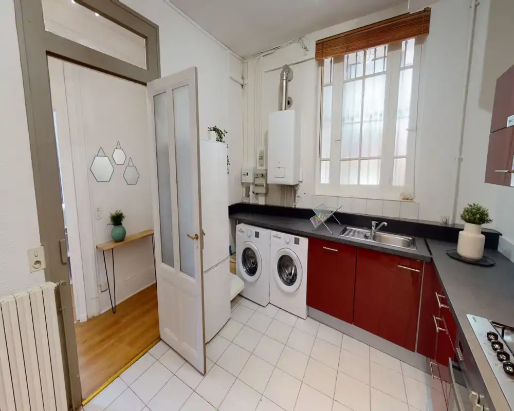 Bright, modern kitchen with red lower cabinets, black countertops, built-in sink and two front-loading laundry machines; large window provides natural light and the space is tidy and functional.