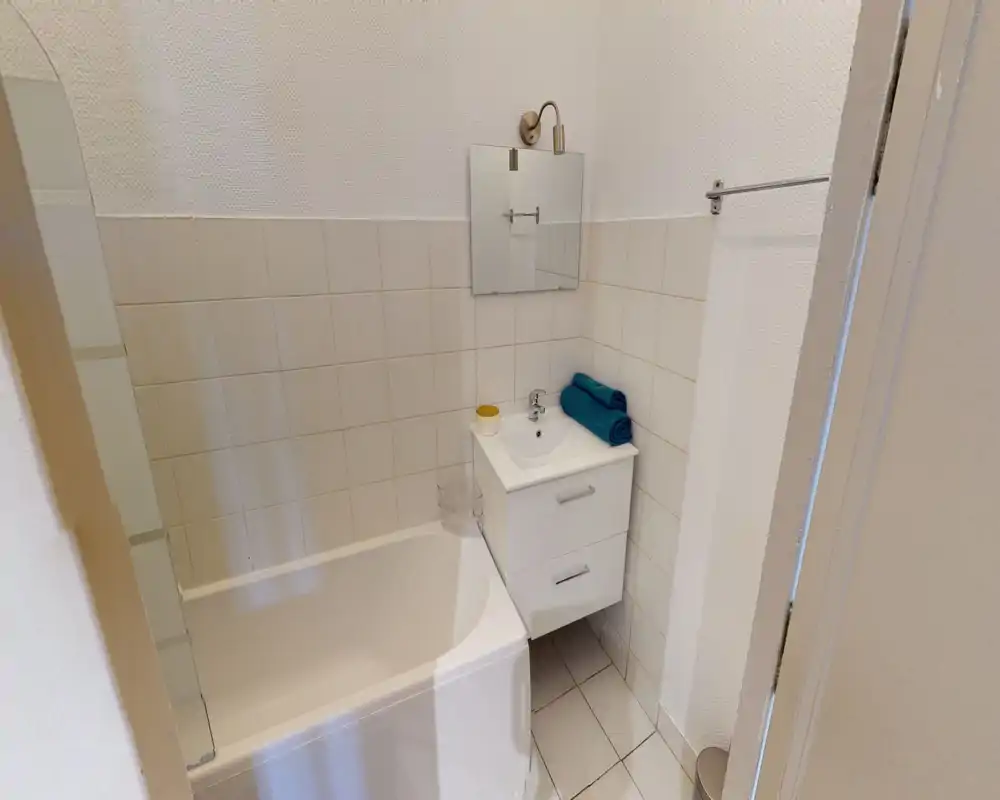 Compact, well-lit bathroom with a bathtub, wall-mounted sink cabinet, mirror and tiled walls. Neat towels add a tidy, functional touch.