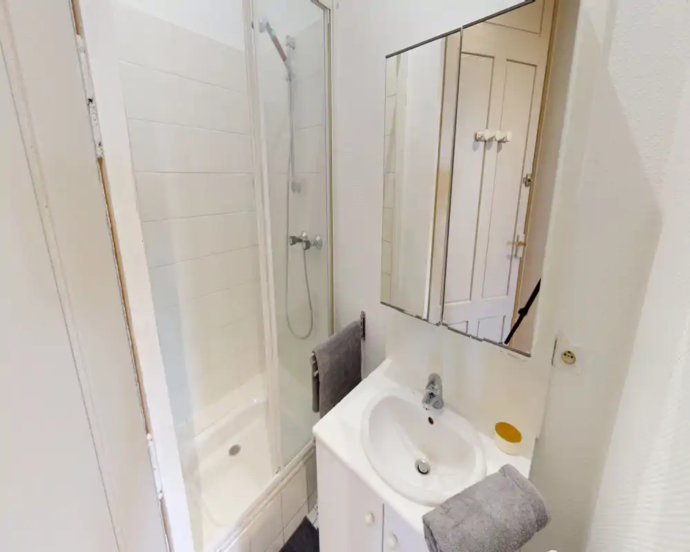 Compact, well-maintained bathroom with a walk-in shower, white tiled walls, a sink with vanity and mirrored cabinet — bright and functional.