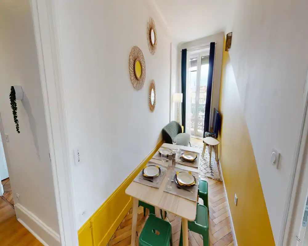 Bright narrow dining area with a small table set for four, green stools and a cozy corner chair by a balcony window — warm yellow accents and decorative wall mirrors add charm.