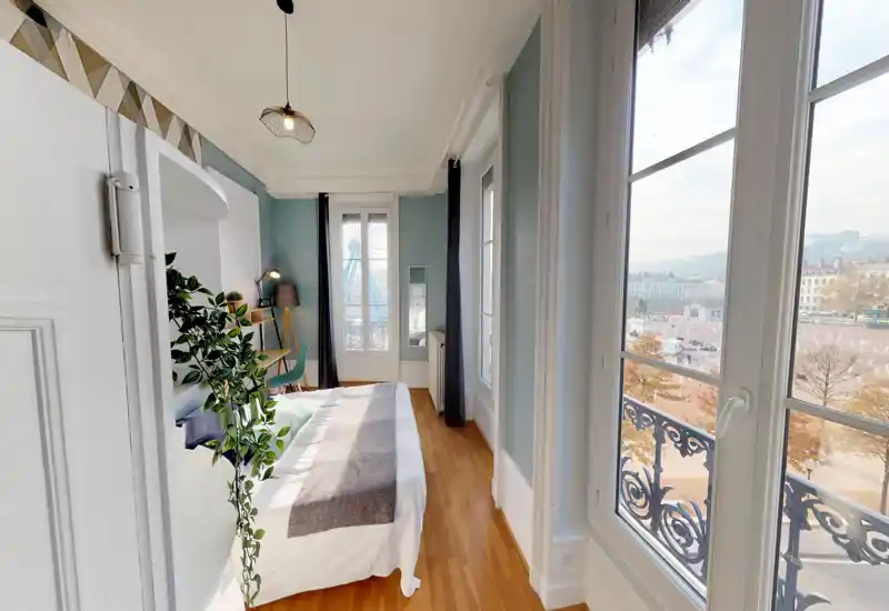 Dormitorio luminoso y con estilo con cama grande, suelo de madera, textiles decorativos y amplia ventana con vistas a la ciudad. Paredes azul claro, rincón de lectura con lámpara y plantas que aportan calidez.