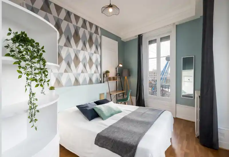 Dormitorio luminoso y con estilo, con una cama doble bien hecha, cojines decorativos y una pared con diseño geométrico. La luz natural entra por una gran ventana y hay un rincón de estudio funcional.