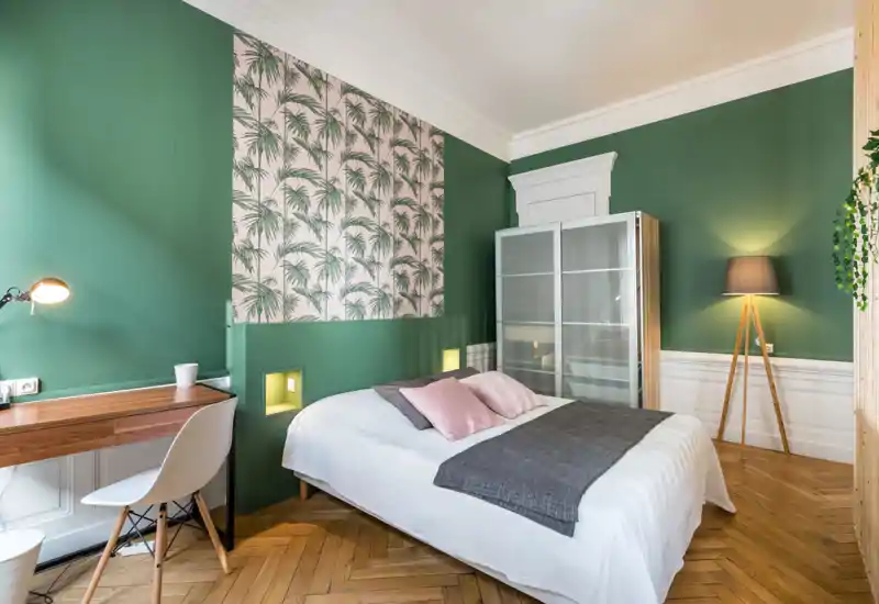 Bright, stylish bedroom with a double bed, pastel pillows and a decorative green-and-pink accent wall. The room includes a wooden desk, modern floor lamp and a wardrobe with frosted sliding doors.