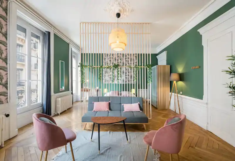 Sunny, stylish living area with a mid-century sofa, two blush armchairs, wooden coffee table and large windows; light green walls and decorative room divider add character.