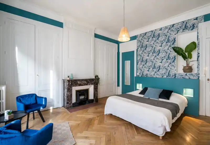 Amplia y elegante habitación con cama doble, pared decorativa y zona de asientos acogedora. Luz natural abundante, suelo de parquet y decoración cuidada que crean un ambiente agradable.
