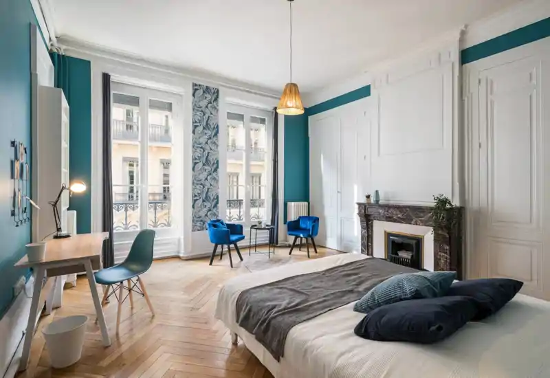 Dormitorio luminoso y elegante con una cama grande en primer plano, suelo de parquet, chimenea decorativa y dos butacas azules junto a las altas ventanas que aportan mucha luz natural.