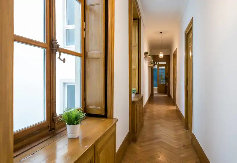 Long couloir lumineux avec parquet et rebord de fenêtre en bois; éclairage chaud et plantes en pot créent une entrée accueillante.
