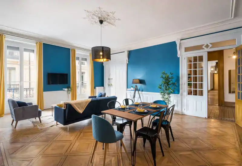Bright, stylish living and dining area with blue accent walls, large windows and elegant parquet flooring — a well-lit, airy space ideal for relaxing and entertaining.