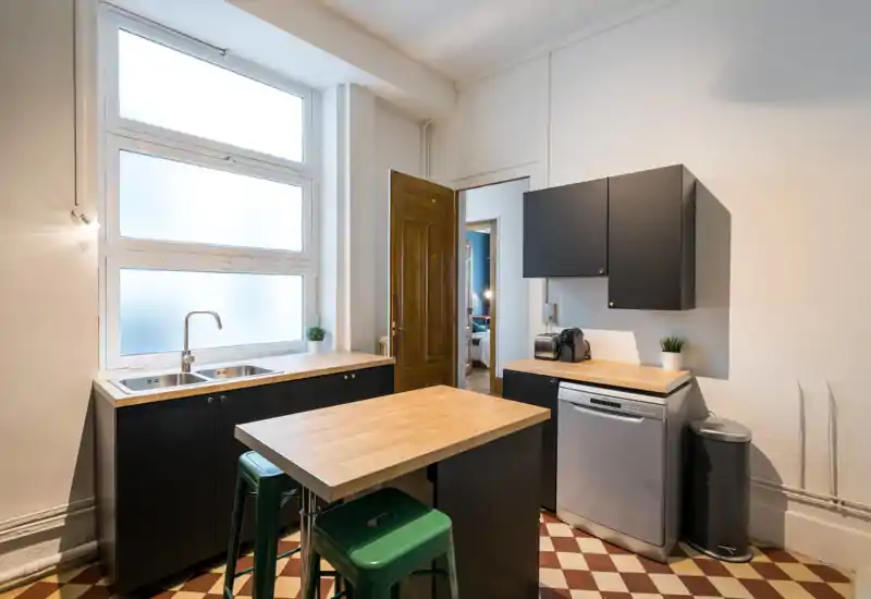 Cuisine moderne et compacte avec plans de travail en bois, placards noirs, évier double sous une large fenêtre dépoli et lave-vaisselle, sur un sol carrelé chaleureux.