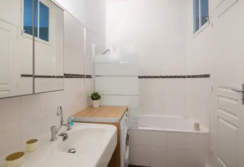Salle de bains propre et lumineuse avec lavabo, armoire miroir, baignoire équipée d'une paroi en verre dépoli et lave-linge placé à côté de la baignoire.