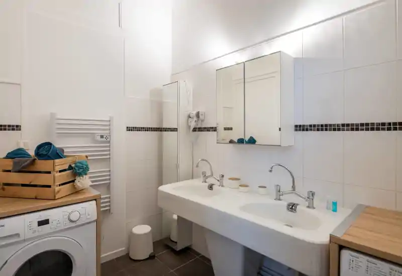 Salle de bains moderne et propre avec vasque double, armoire miroir et carrelage blanc. Chauffe-serviettes, sèche-cheveux et emplacement lave-linge visibles, pratique pour un séjour confortable.
