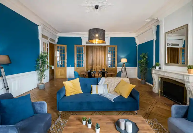 Grand salon élégant aux murs bleu profond, canapés en velours et coussins jaunes; moulures raffinées et cheminée en marbre pour un charme classique.