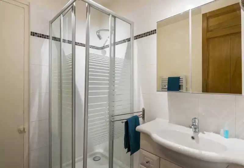 Salle de bains propre et compacte avec douche vitrée, lavabo blanc et armoire miroir. Bien éclairée et présentée avec soin — parfaite pour les locations courte durée.