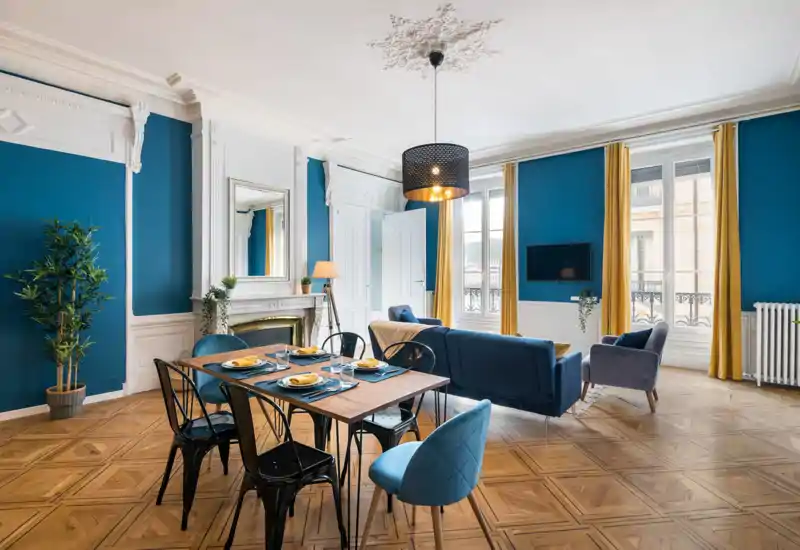 Grand salon avec coin repas élégant : murs bleu profond, canapé confortable, cheminée décorative et table dressée — la lumière naturelle des grandes fenêtres crée une ambiance accueillante.