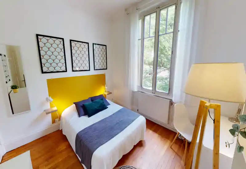 Bright, stylish bedroom with a double bed, bold yellow headboard and large window letting in abundant natural light. The room features warm wooden floors, decorative pillows and minimal modern furnishings for a cozy, contemporary feel.