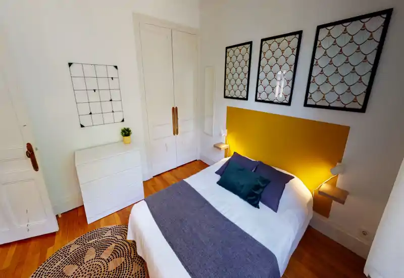 Sunny, well-styled bedroom with a double bed, bold mustard headboard and decorative framed art. Hardwood floors, soft linens and bedside lamps create a cozy, welcoming atmosphere ideal for short-term rentals.