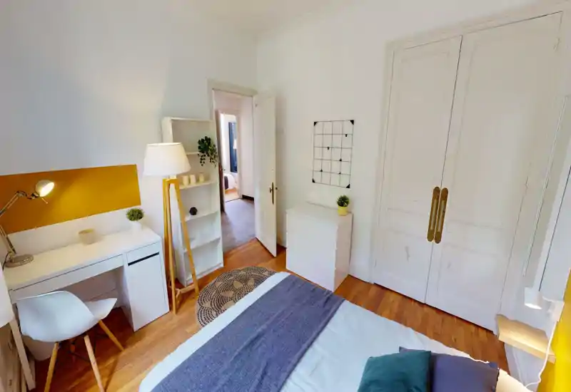 Bright, cozy bedroom with a comfortable bed in the foreground, wooden floors, a small desk and shelving, and soft decorative lighting — ideal for a restful stay.