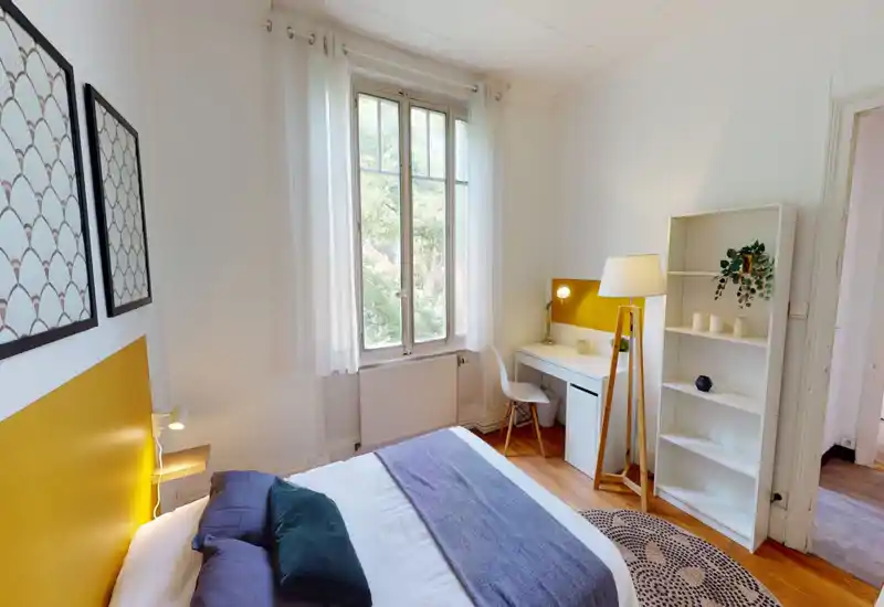 Bright, stylish bedroom with a large central bed, soft textiles and a desk by the window. Natural light and warm yellow accents create an inviting atmosphere.
