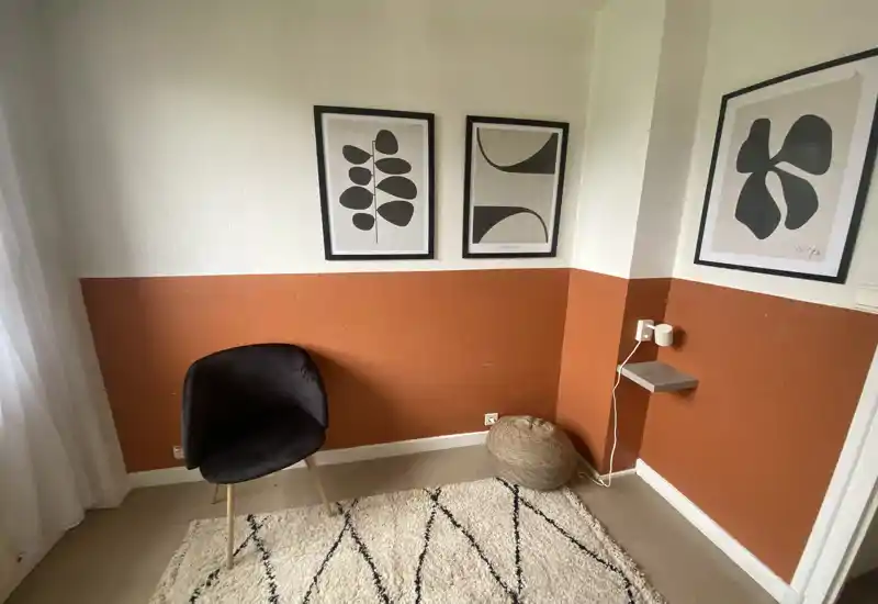 Cozy living corner with an accent chair, woven pouf and decorative rug against a warm two-tone wall with framed abstract prints — a stylish spot for reading or relaxing.