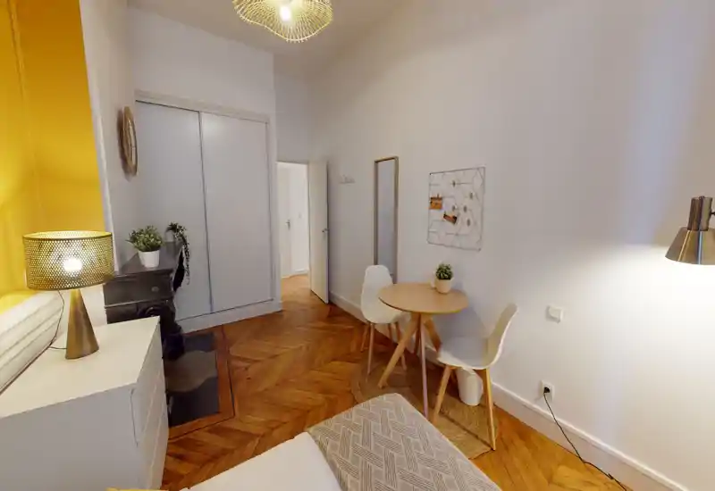 Cozy, well-lit bedroom corner with a visible bed in the foreground, warm wooden herringbone floors, a small round dining table with two chairs, and decorative plants and lighting creating a welcoming atmosphere.