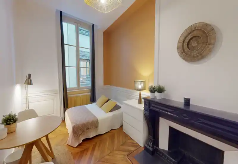 Sunny, well-styled bedroom with a cozy bed, warm accent wall and decorative fireplace; natural light from a large window enhances the wooden herringbone floor and modern furnishings.