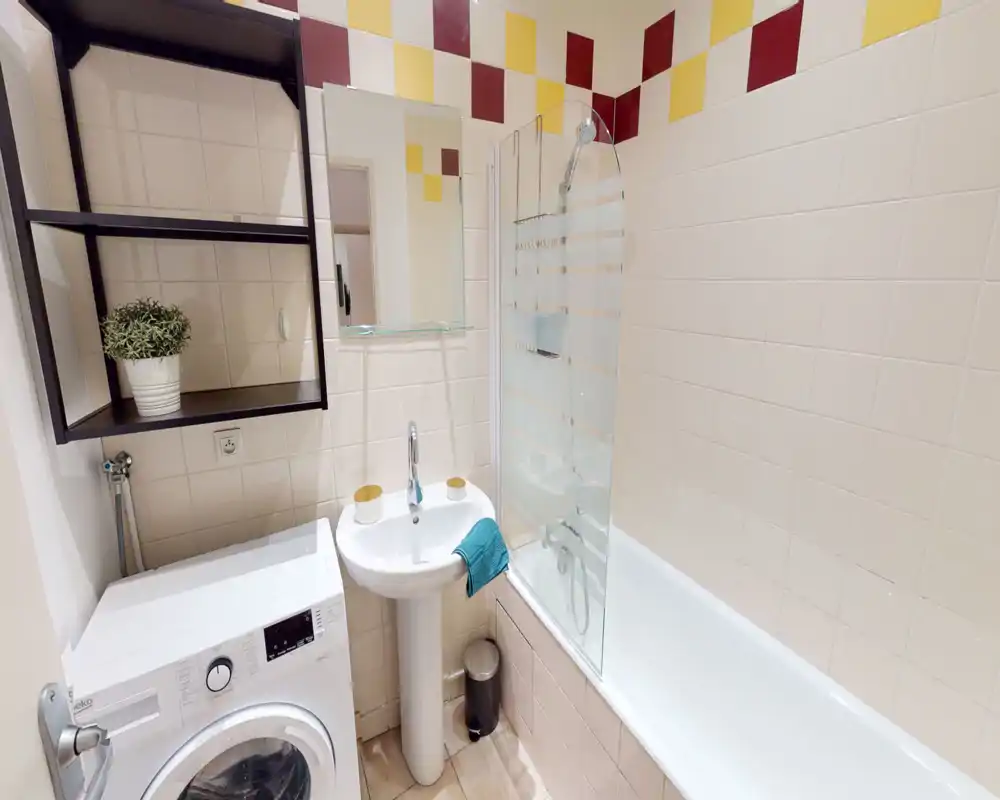 Compact, clean bathroom with a bathtub and glass shower screen, pedestal sink, washing machine and modern tilework with colorful accents.