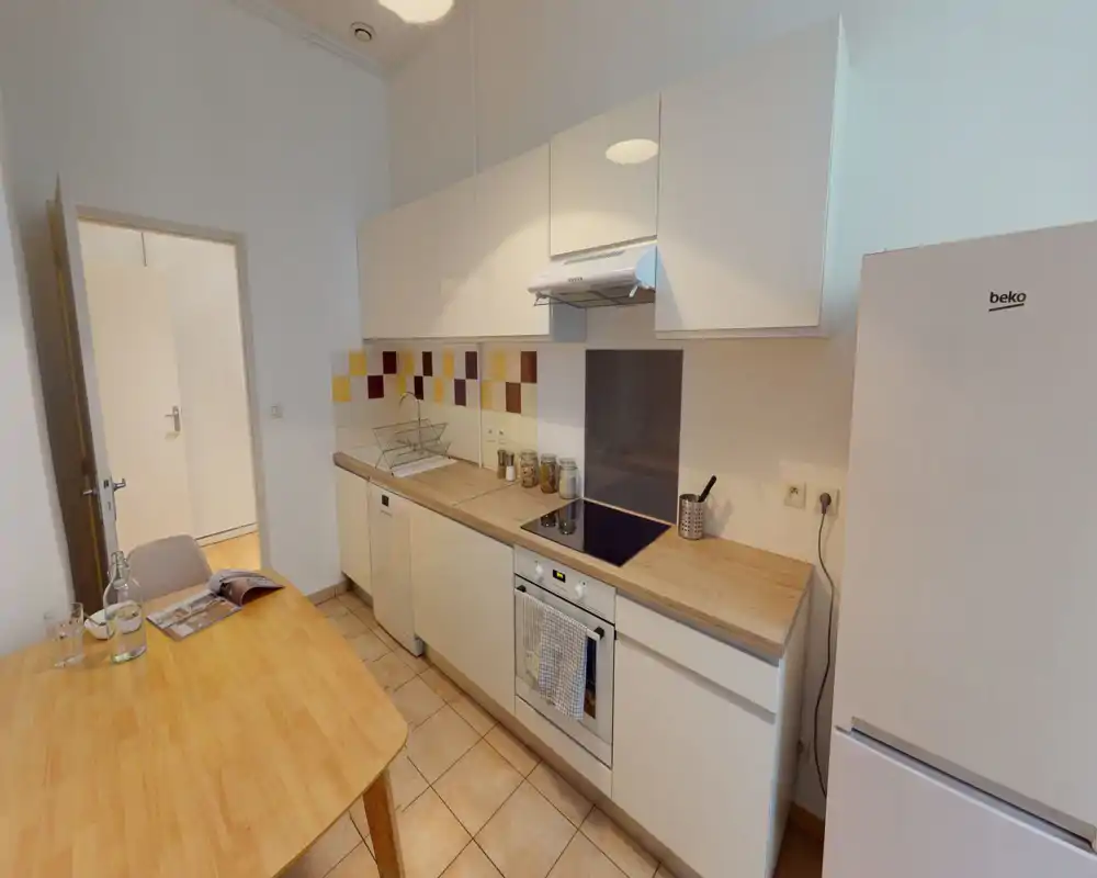 Bright, modern kitchenette with white cabinets, wooden countertops and a dining table. Appliances include an oven, stovetop, dishwasher and a refrigerator, plus practical storage jars and a dish rack.