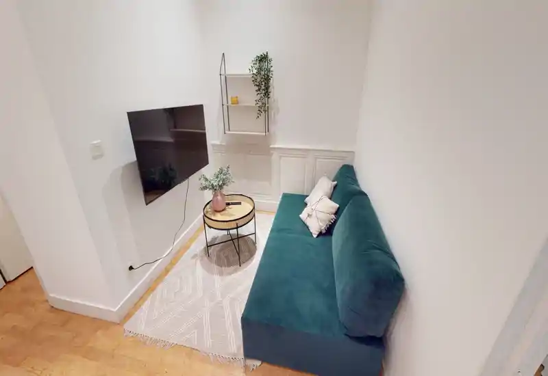 Compact, stylish living area with a teal sofa, decorative cushions, a small round coffee table and wall-mounted TV — bright, cozy and well styled for a modern rental.