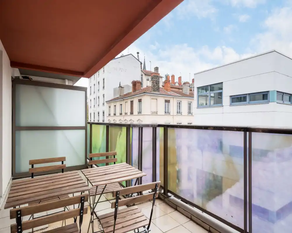 Balcone cittadino soleggiato con tavolo e sedie in legno in stile bistro, pannelli di vetro colorati semi-opachi e vista sui tetti circostanti — perfetto per il caffè del mattino.