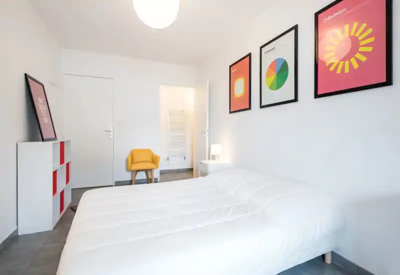 Camera da letto luminosa e minimale con letto ben rifatto, quadri moderni e una poltroncina gialla. Linee pulite e luce soffusa creano un ambiente tranquillo e accogliente.