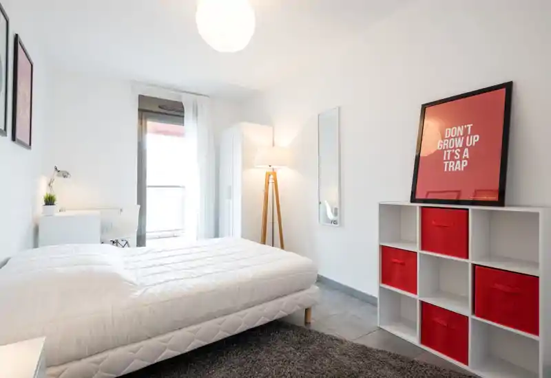 Camera da letto moderna e luminosa con un letto matrimoniale completamente visibile, biancheria bianca, una scrivania vicino alla finestra e scaffalatura con contenitori rossi: pulita, ben illuminata e ben arredata.