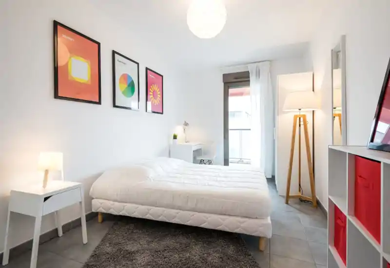 Camera da letto moderna e luminosa con ampio letto bianco, comodino e scrivania vicino alla finestra. Arredamento minimal e illuminazione calda rendono l’ambiente accogliente, perfetto per annunci.