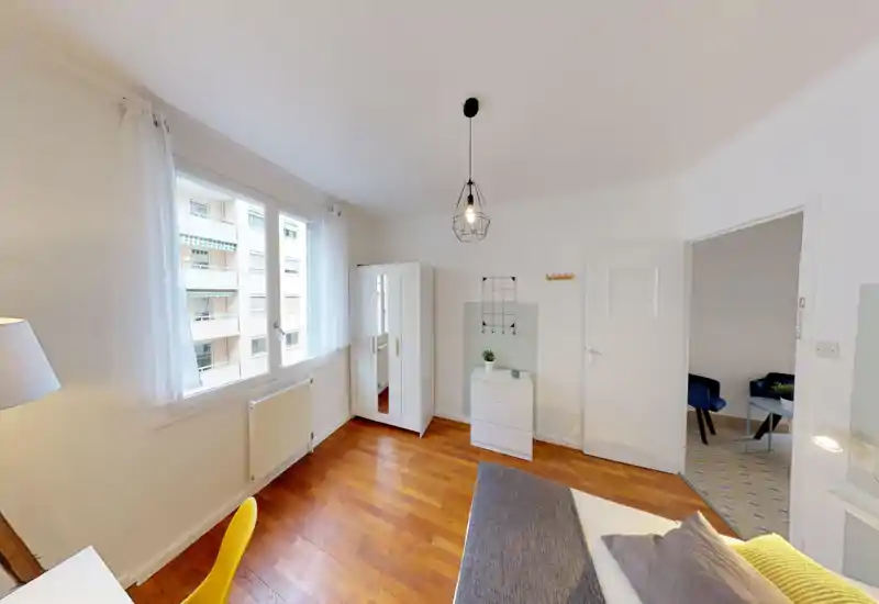 Bright, simply furnished bedroom with wooden floors, a white wardrobe and a partially visible bed with cushions and a throw; large windows provide ample natural light.