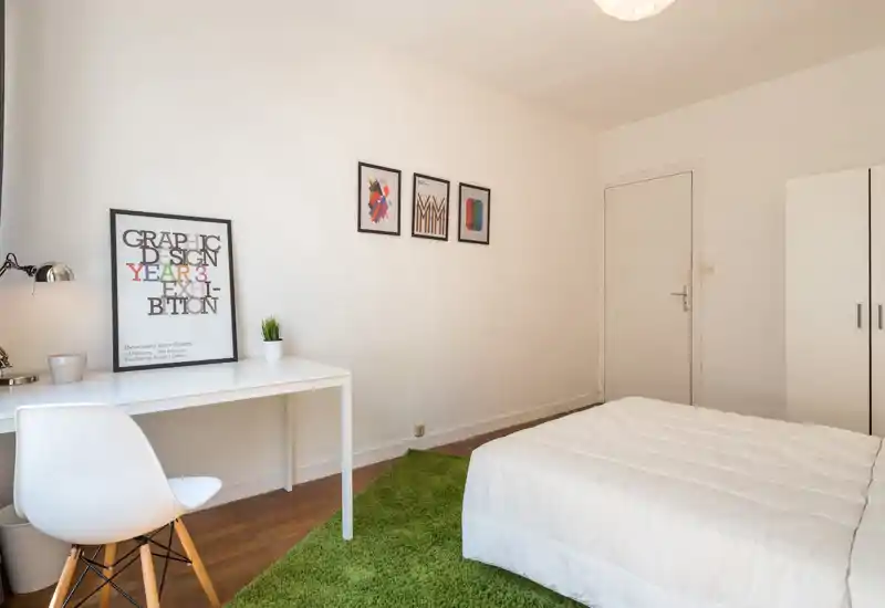 Bright, minimal bedroom with a white bed, green rug and a tidy desk — ideal for a calm, modern rental. Simple wall art and natural light make the room feel airy and welcoming.