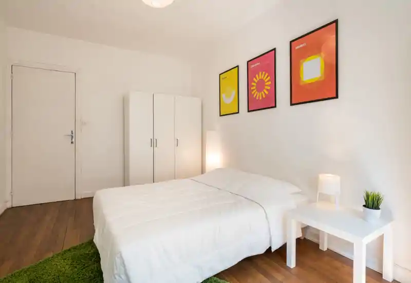 Bright, minimalist bedroom with a neatly made double bed, white wardrobe, bedside table with lamp and framed colorful prints on the wall — calm, modern styling ideal for rental listings.