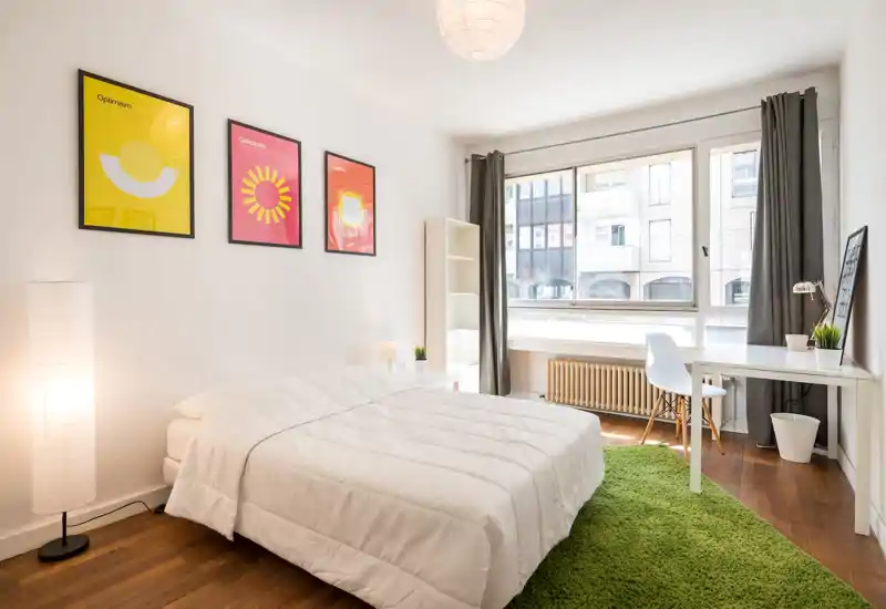 Bright, modern bedroom with a large bed dressed in white linens, a green rug, desk by the window and colorful wall art—well lit by a big window providing pleasant natural light.