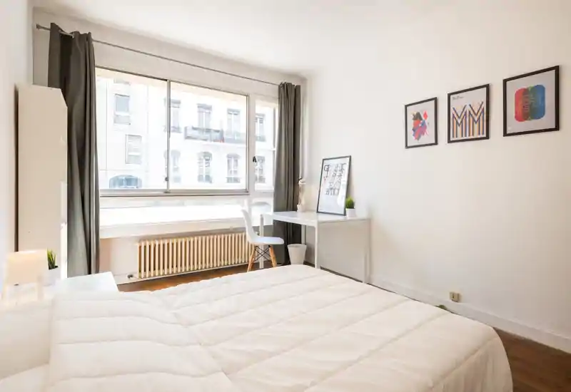Bright, minimalist bedroom with a large window, white bedding and a small work desk — ideal for a calm, sunny stay.