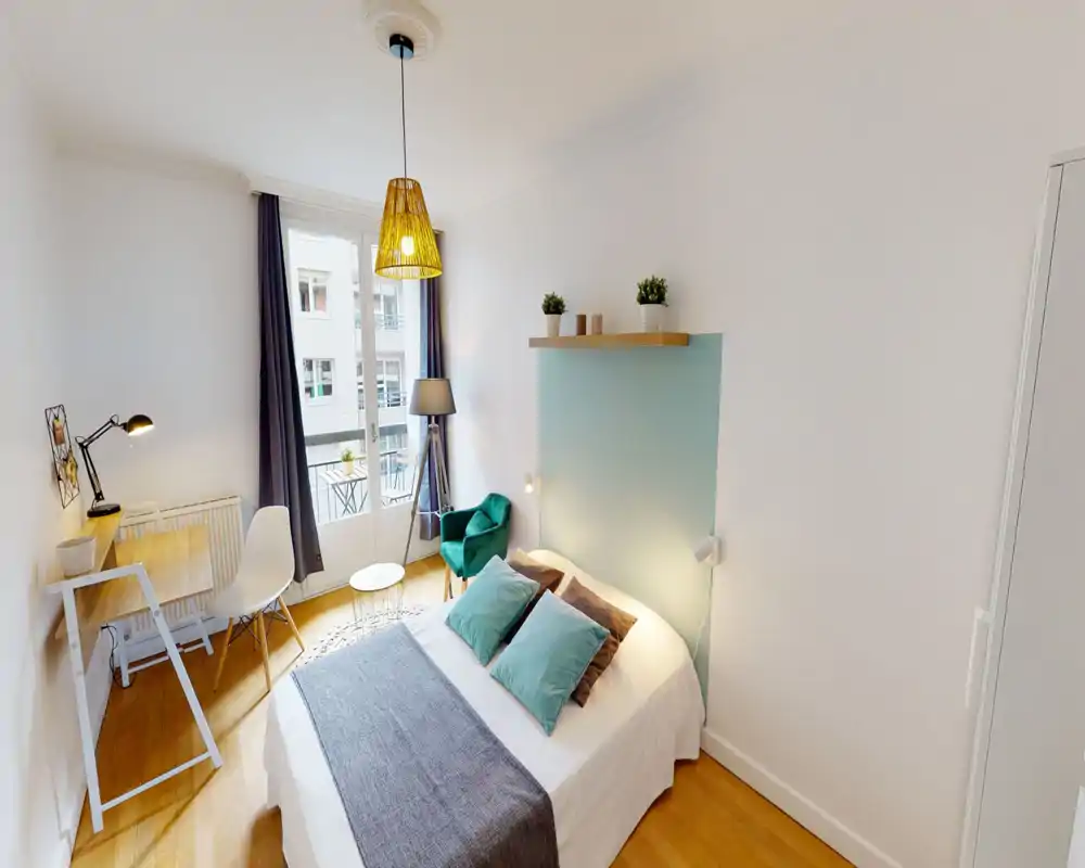 Dormitorio moderno y luminoso con cama doble cómoda, cojines decorativos y un sillón acogedor junto a la ventana. Un escritorio pequeño y puertas al balcón aportan luz natural y un agradable espacio de trabajo.